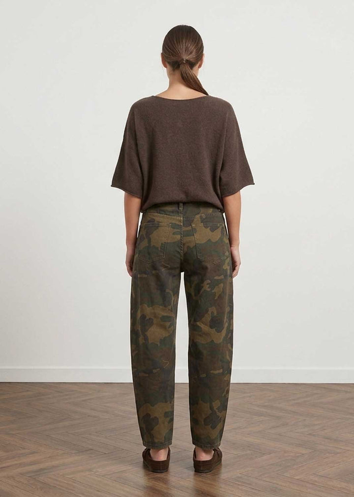 A woman viewed from behind wearing a dark brown short-sleeve top and camouflage pants, standing on a wood floor against a plain white wall.