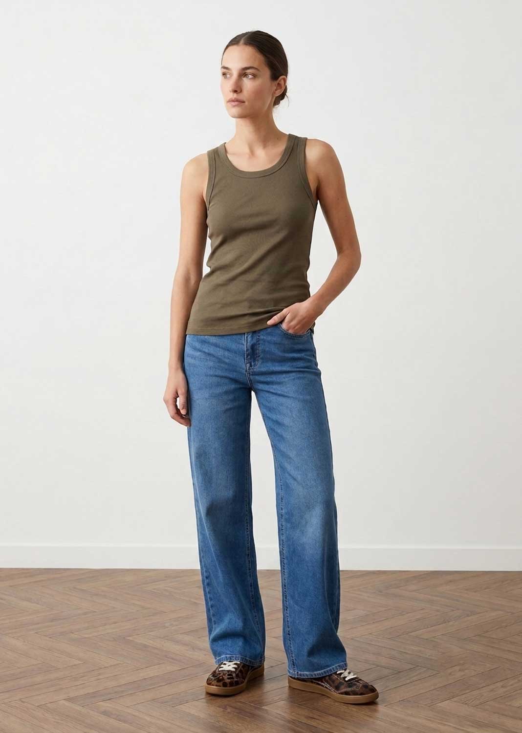 A woman modeling a sleeveless olive green tank top and wide-leg blue jeans, standing on a wood herringbone floor against a plain white wall.