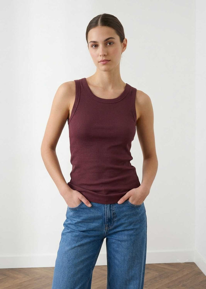 A woman modeling a sleeveless crew-neck tank top in deep burgundy, paired with blue jeans, standing against a plain white wall.
