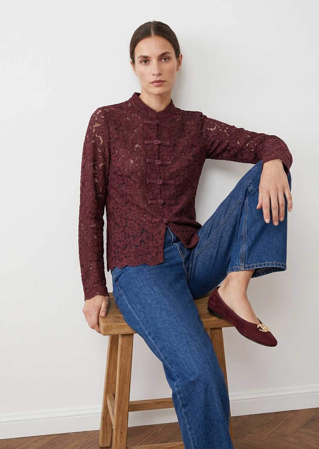 A woman models a burgundy lace long-sleeve blouse with a mandarin collar and front buttons, paired with blue jeans and burgundy flats, sitting on a wooden stool against a white wall.