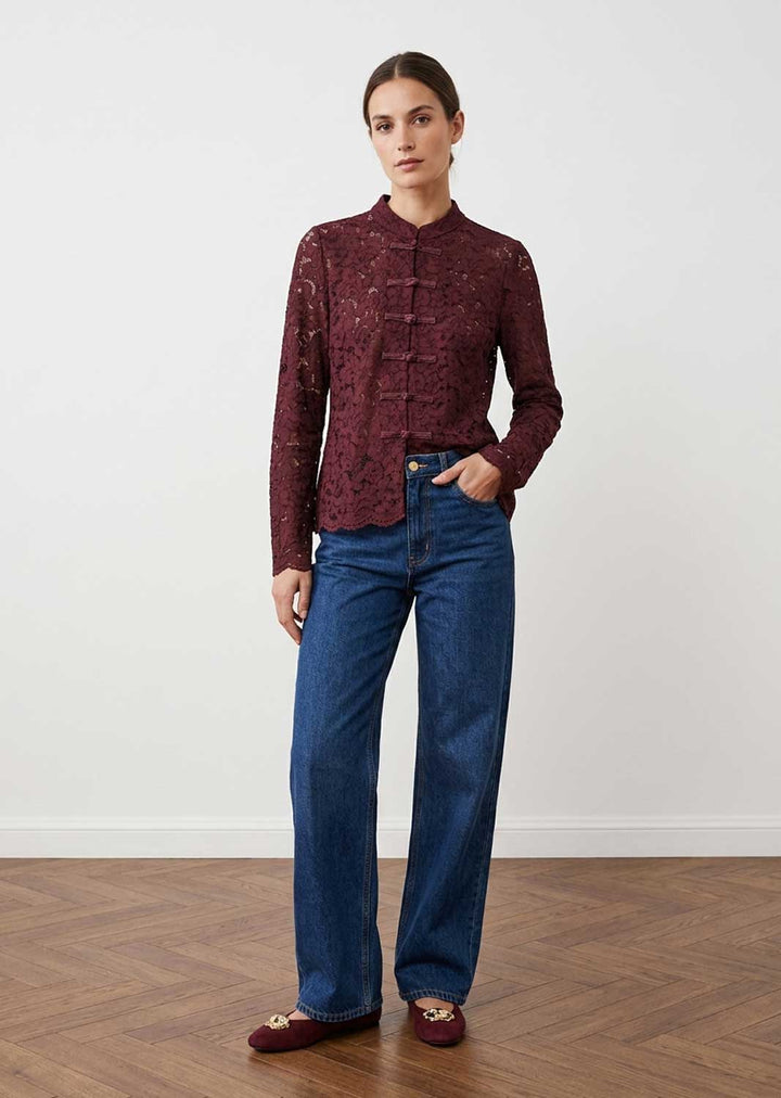 A woman modeling a long-sleeve burgundy lace blouse with a mandarin collar and button placket, paired with blue jeans and burgundy flats, standing against a white wall and wood floor.