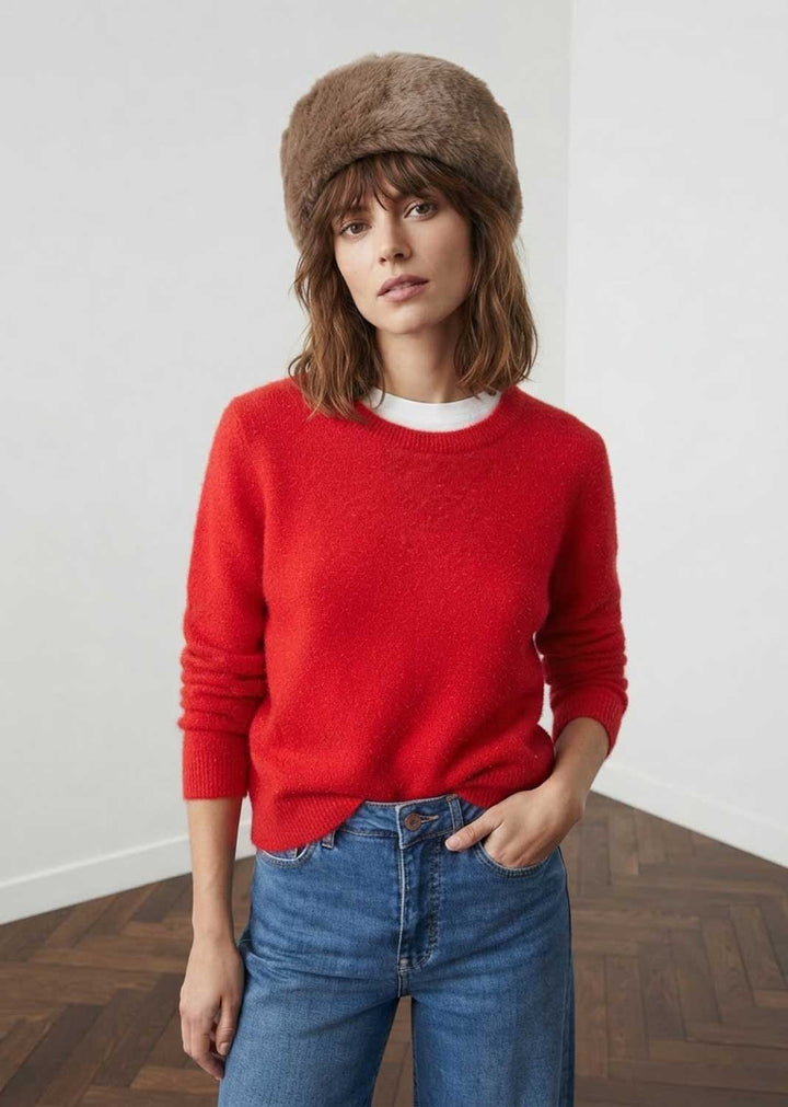 A woman wearing a bright red knit crewneck sweater, blue jeans, and a brown faux fur cossack hat, standing in a minimalist room with white walls and a wooden parquet floor.