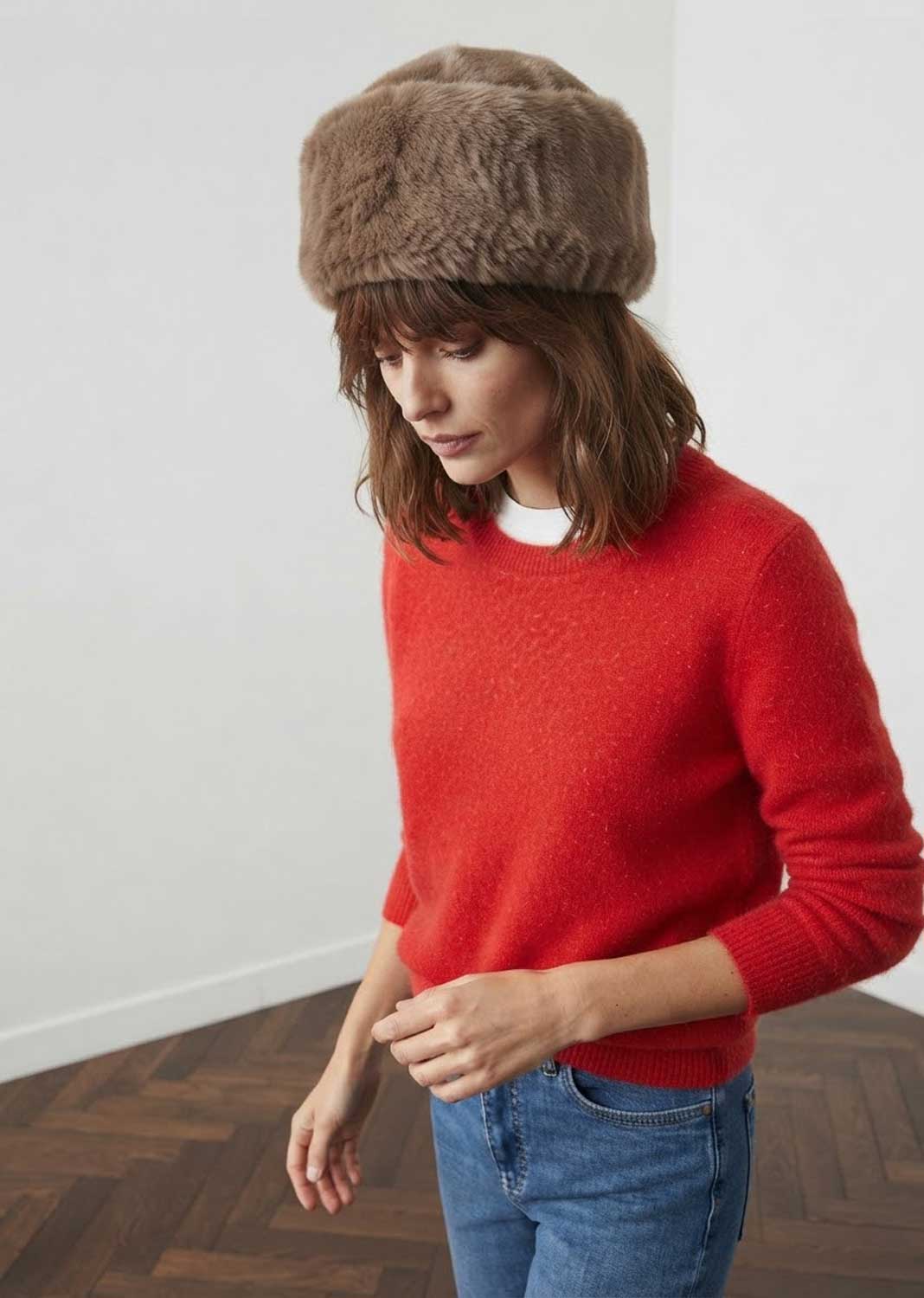 A woman wearing a brown faux fur cossack hat, a red knit sweater, and blue jeans, standing in a minimalist room with a white wall and wood parquet floor.