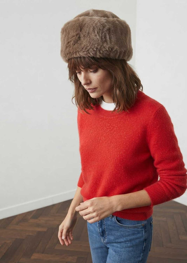 A woman wearing a brown faux fur cossack hat, a red knit sweater, and blue jeans, standing in a minimalist room with a white wall and wood parquet floor.