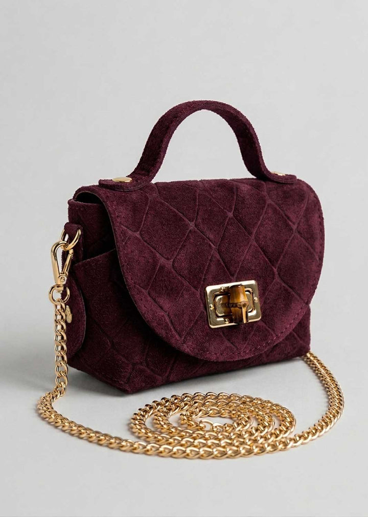 A burgundy croc suede handbag with a top handle, gold-tone clasp, and a long gold chain strap, displayed on a light gray background.