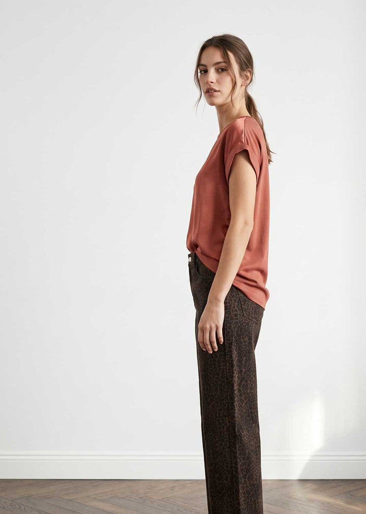 A woman stands in profile wearing a muted terracotta short-sleeve top and dark brown patterned wide-leg trousers against a plain white wall.