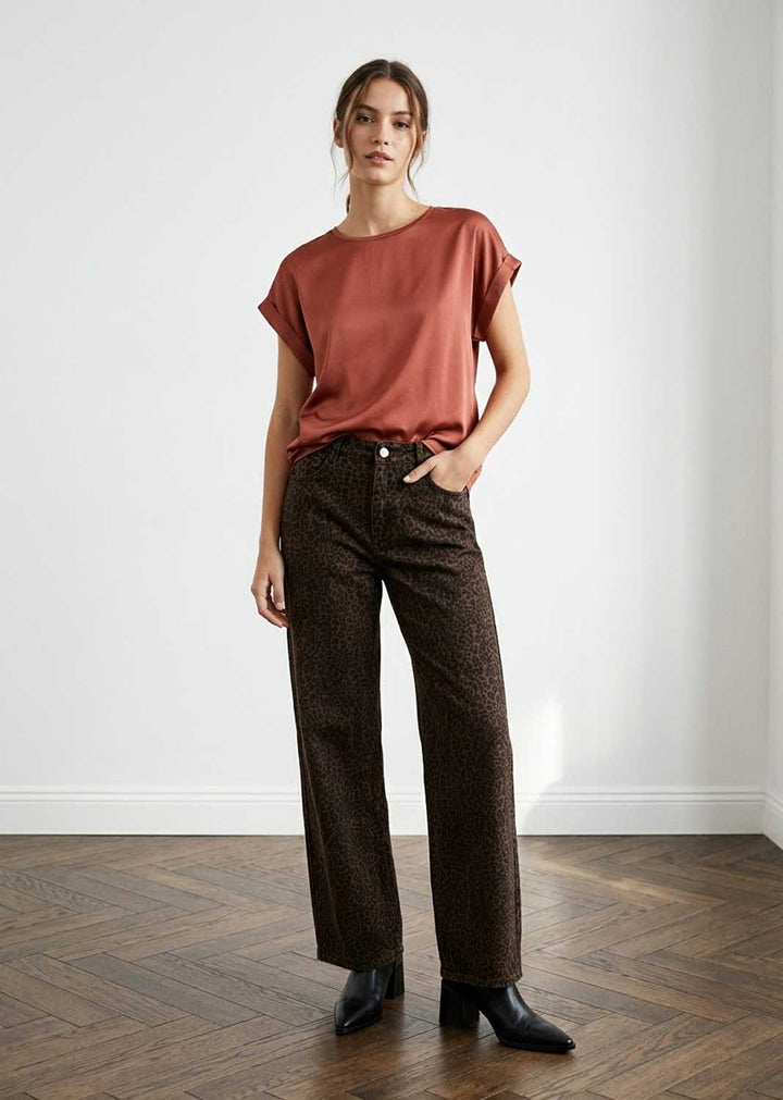 A woman stands in a minimalist room with a white wall and wood parquet floor, wearing a rust-colored short-sleeve top, dark brown patterned wide-leg trousers, and black ankle boots.