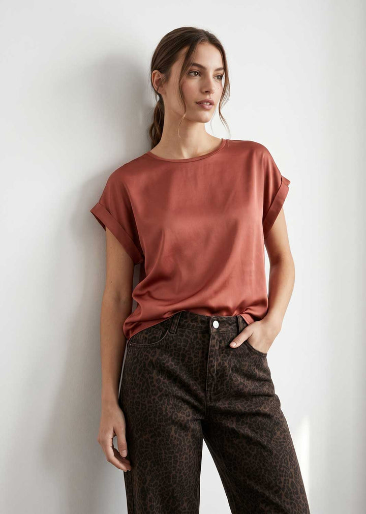 A woman wearing a rust-colored short-sleeve top and dark brown leopard-print pants, standing against a plain light gray wall.