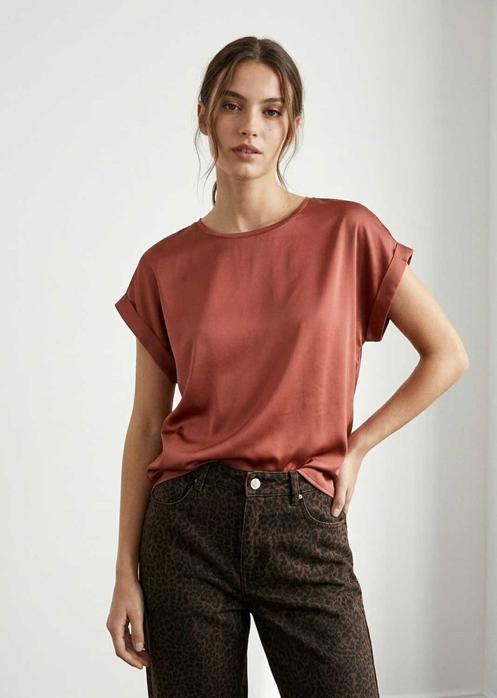 A woman modeling a short-sleeve, crew-neck satin top in a muted terracotta or rust color, paired with dark brown patterned jeans, against a plain light gray studio background.