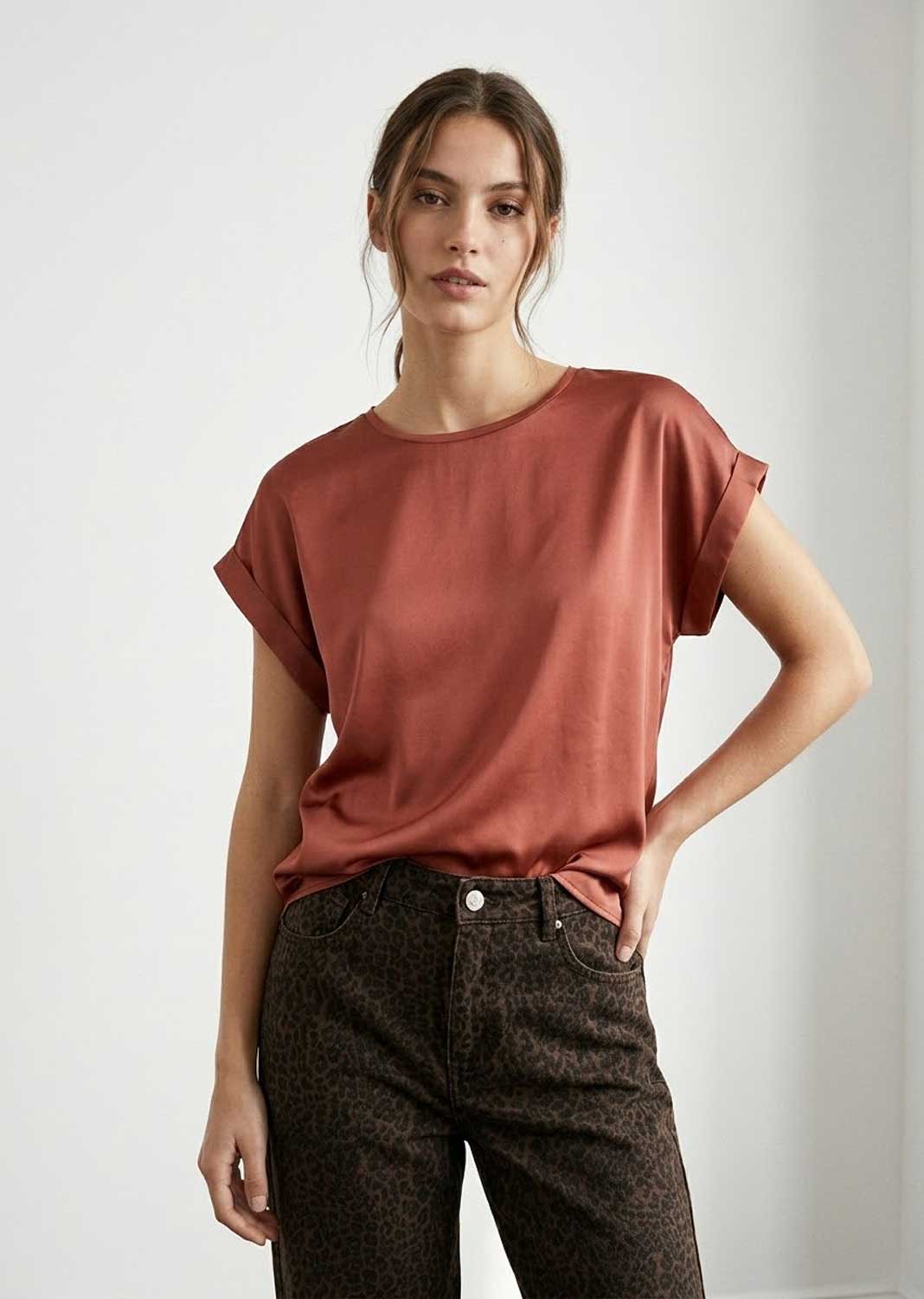 A woman modeling a short-sleeve, crew-neck satin top in a muted terracotta or rust color, paired with dark brown patterned jeans, against a plain light gray studio background.