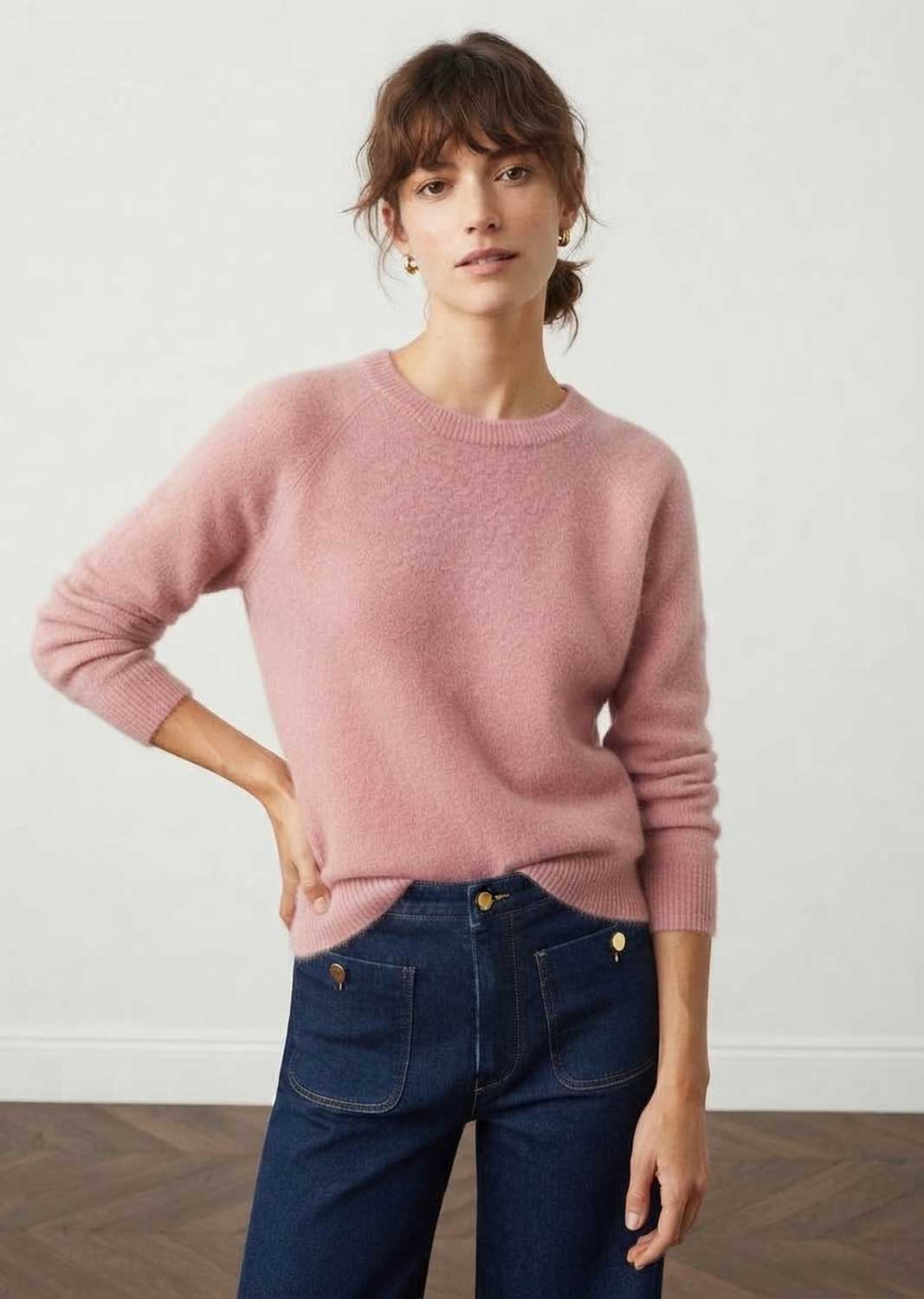 A woman wearing a dusty pink crewneck knit sweater and dark blue jeans, standing against a plain off-white wall.