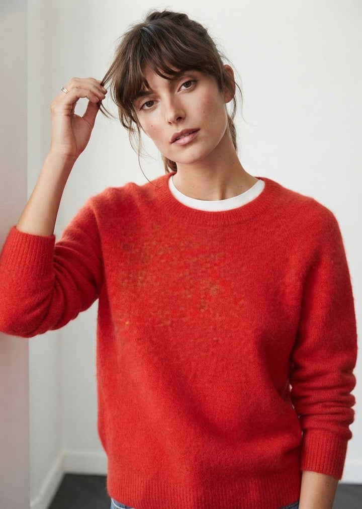 A woman wearing a bright red fuzzy crewneck sweater over a white undershirt, standing against a plain light wall.