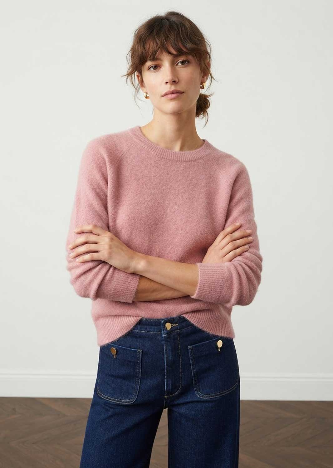 A woman wearing a dusty pink knit crewneck sweater and dark blue jeans, standing with arms crossed against a plain white wall.