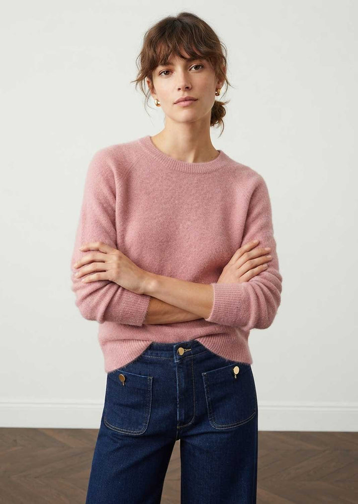 A woman wearing a dusty pink knit crewneck sweater and dark blue jeans, standing with arms crossed against a plain white wall.