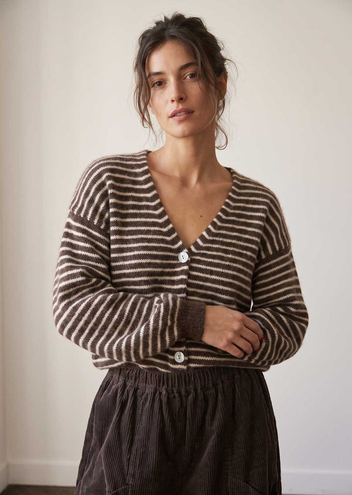 A woman wearing a brown and beige striped knit cardigan with white buttons, paired with a dark brown corduroy skirt, standing against a plain off-white wall.