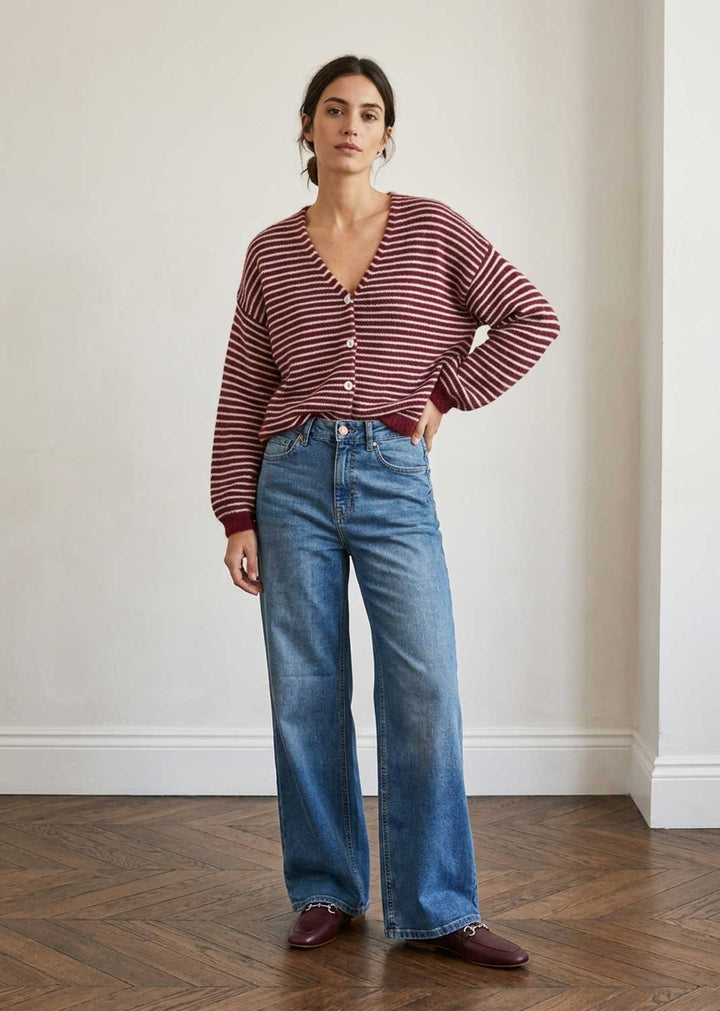 A woman stands in a room with a light beige wall and wooden floor, wearing a burgundy and white striped long-sleeve button-up cardigan, blue wide-leg jeans, and burgundy loafers.