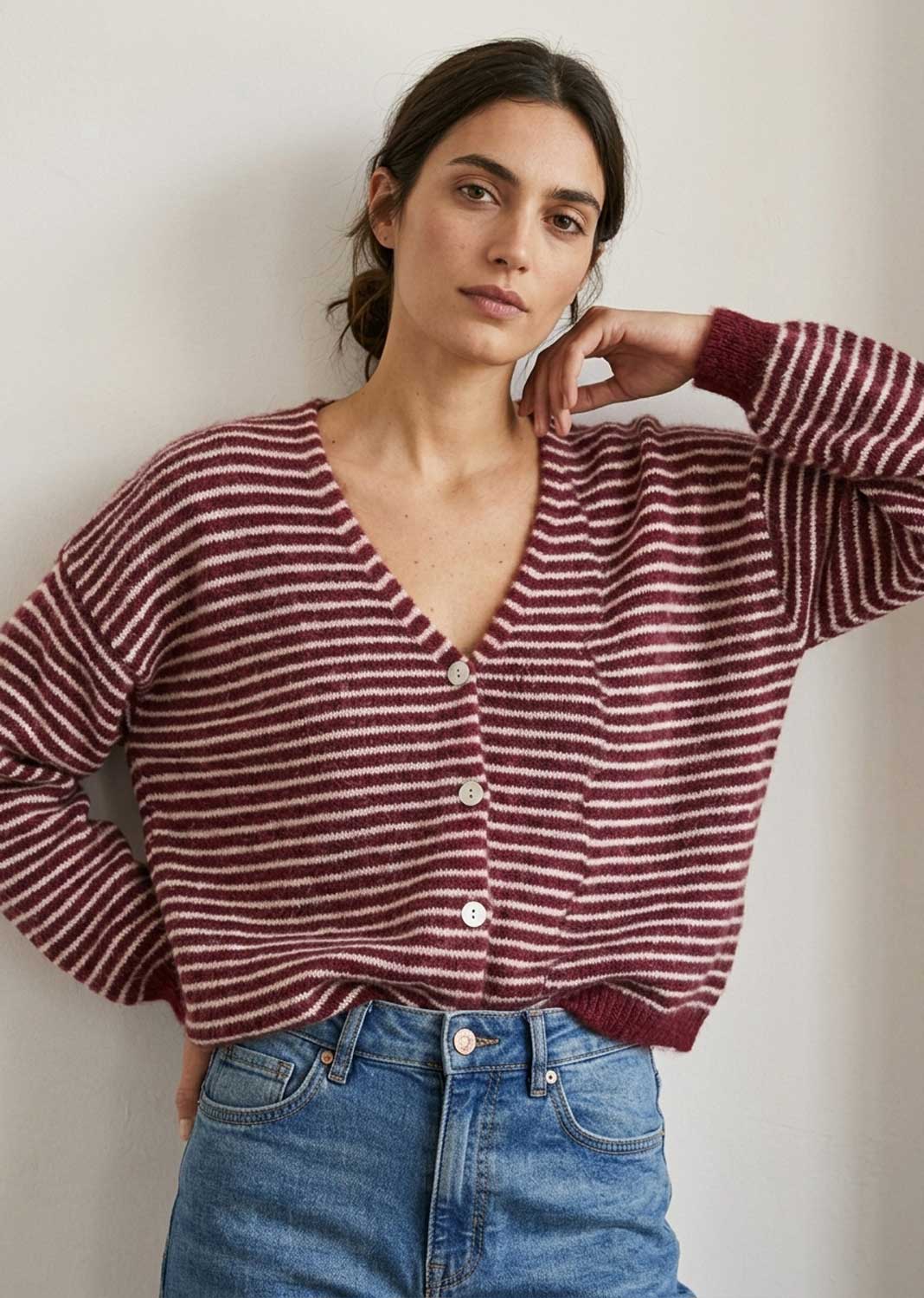 A woman wearing a burgundy and white striped knit cardigan with silver buttons, paired with blue jeans, posing against a neutral off-white wall.
