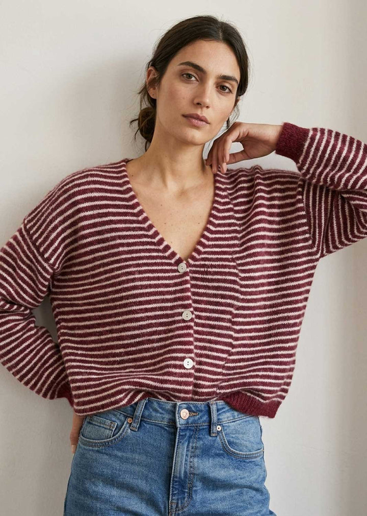 A woman wearing a burgundy and white striped knit cardigan with silver buttons, paired with blue jeans, posing against a neutral off-white wall.
