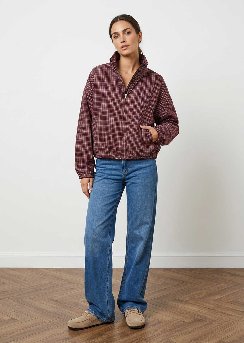 A woman stands in a studio wearing a burgundy and black checkered quarter-zip pullover, blue wide-leg jeans, and beige shoes against a white wall and wood floor.