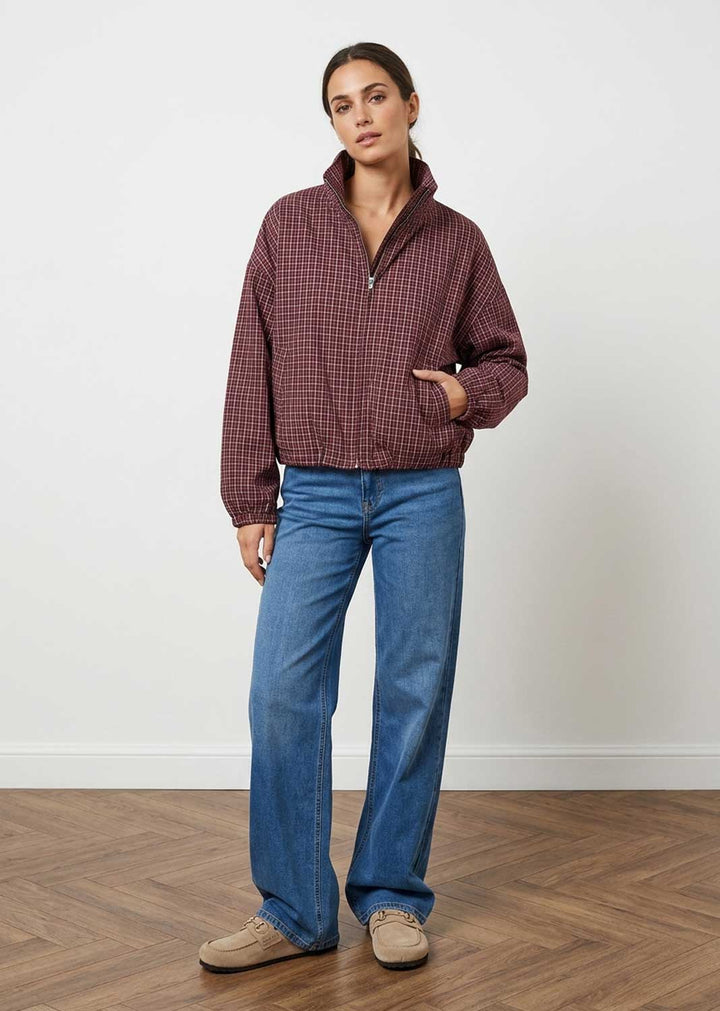 A woman stands in a studio wearing a burgundy and black checkered quarter-zip pullover, blue wide-leg jeans, and beige shoes against a white wall and wood floor.