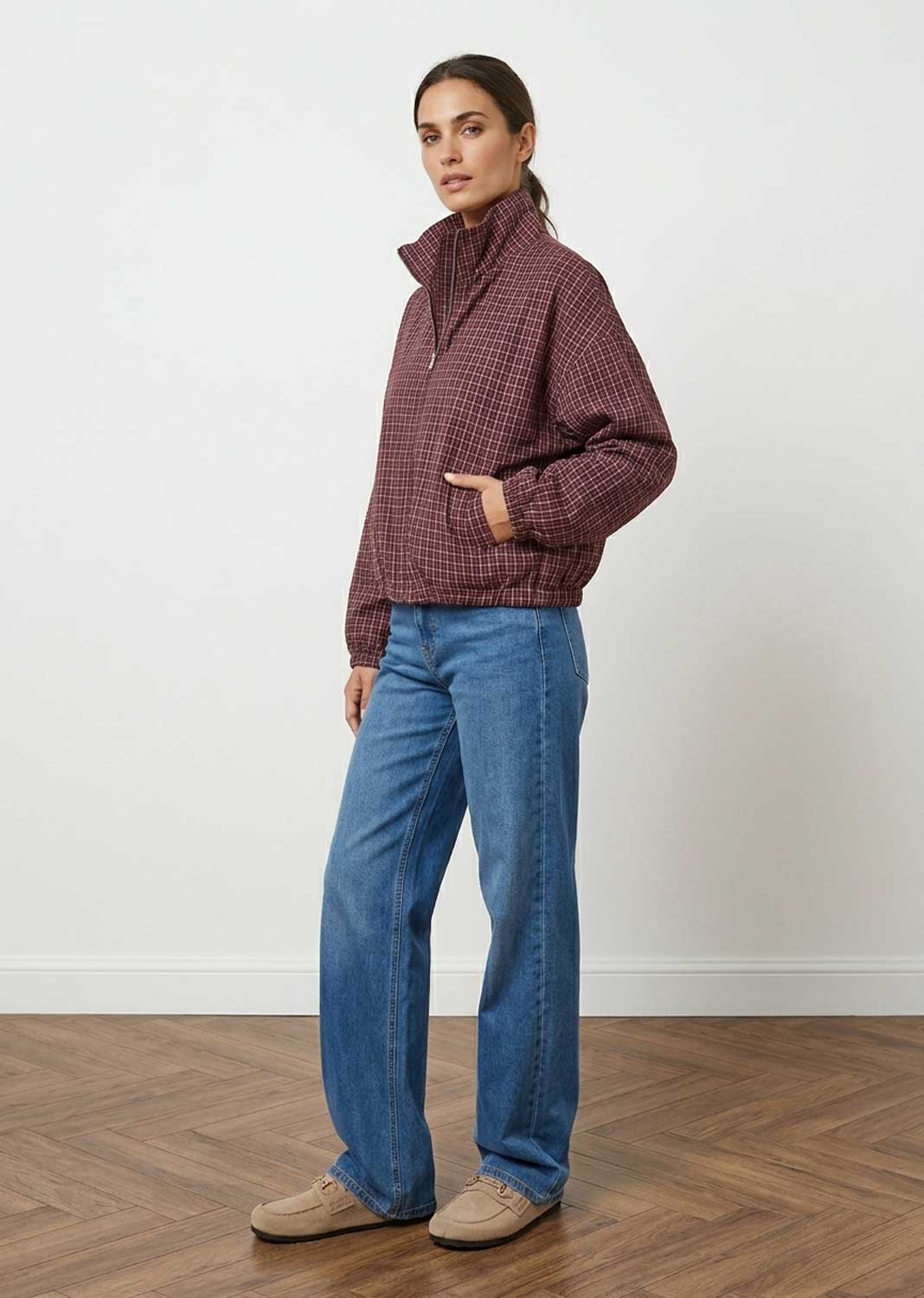 A woman modeling a dark red and black checkered quarter-zip jacket, paired with blue jeans and beige shoes, standing in a room with a white wall and wooden floor.