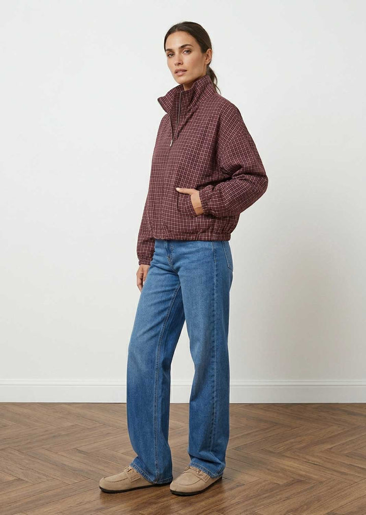 A woman modeling a dark red and black checkered quarter-zip jacket, paired with blue jeans and beige shoes, standing in a room with a white wall and wooden floor.