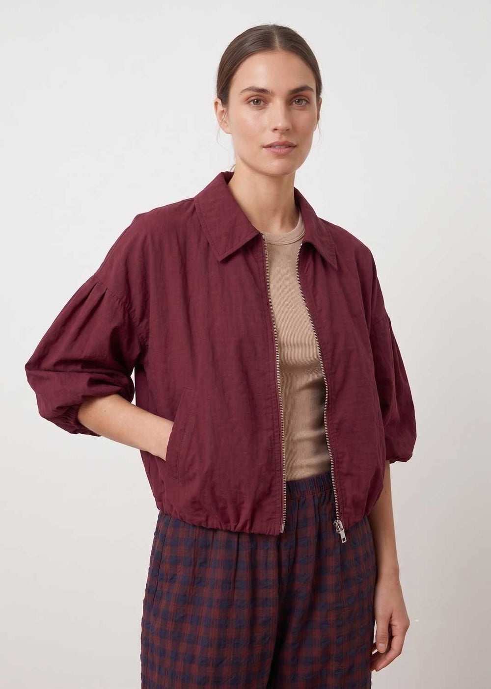 A woman modeling a cropped, oversized button-up jacket in deep burgundy with voluminous sleeves, worn over a beige top and paired with dark blue plaid trousers, against a plain light gray background.