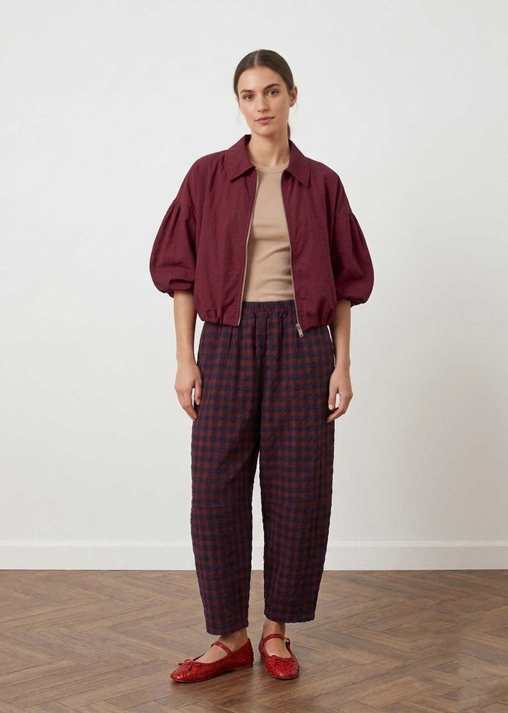 A model stands in a studio wearing a burgundy cropped jacket over a beige top, paired with dark red and navy plaid cropped trousers and red flats, against a white wall and wood floor.