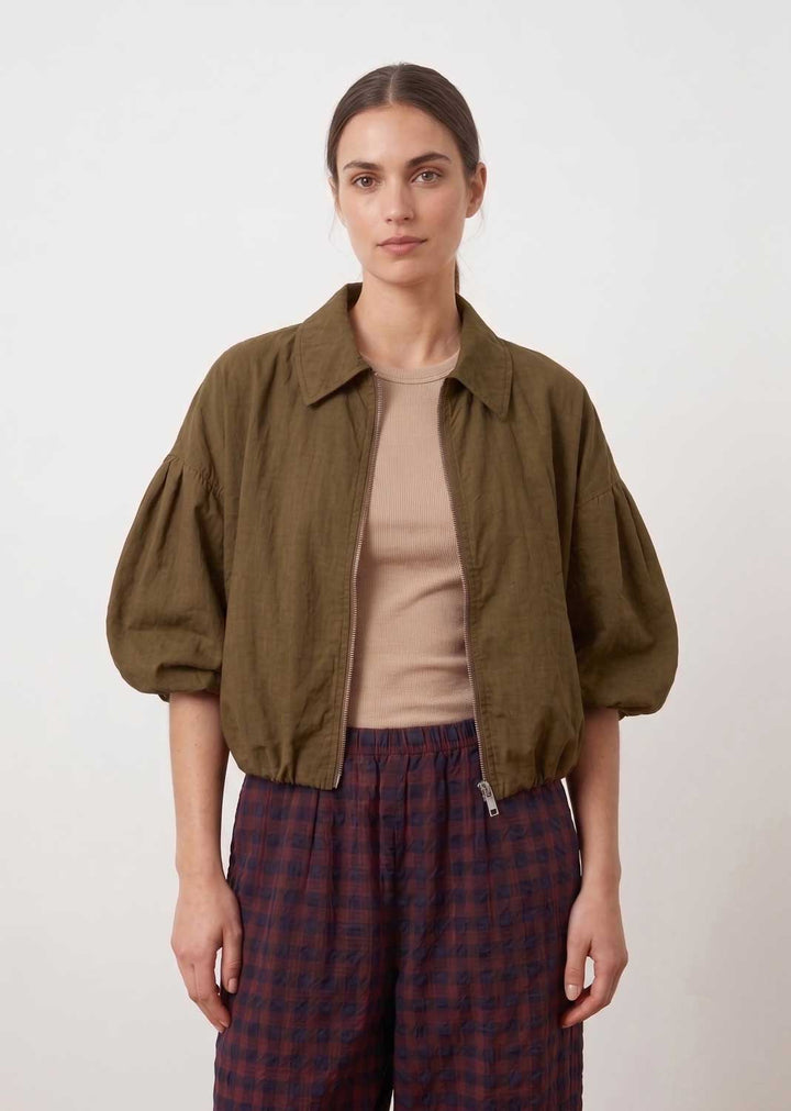 A woman modeling a cropped olive green jacket with voluminous puff sleeves, worn over a beige top and paired with dark red and navy plaid pants, against a plain light gray background.