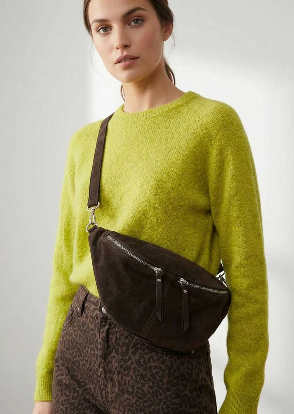 A model wearing a bright lime green knit sweater and brown leopard-print trousers, carrying a dark brown suede crossbody bag with silver hardware.