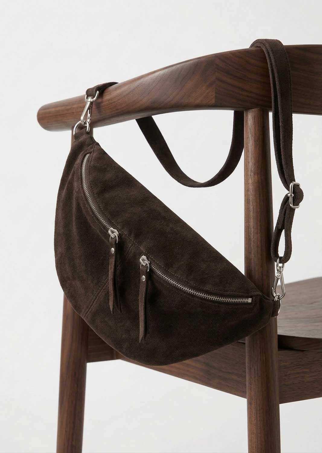 A dark brown suede crescent-shaped shoulder bag with silver zippers and leather straps, hanging from a wooden chair against a light gray background.