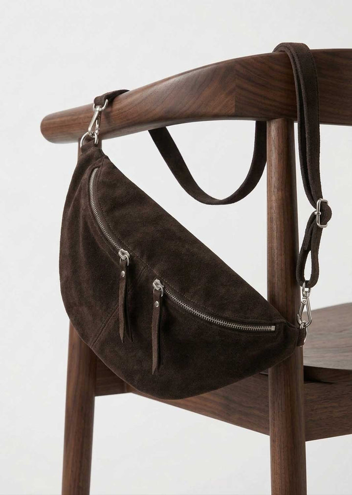 A dark brown suede crescent-shaped shoulder bag with silver zippers and leather straps, hanging from a wooden chair against a light gray background.