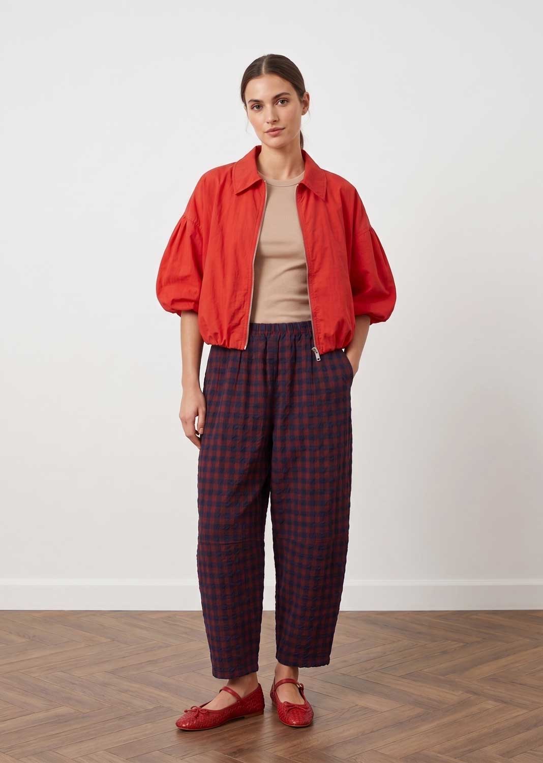 A woman stands in a studio wearing a bright red cropped jacket over a beige top, with dark red and navy plaid cropped pants and red flats, against a plain light wall and wood floor.