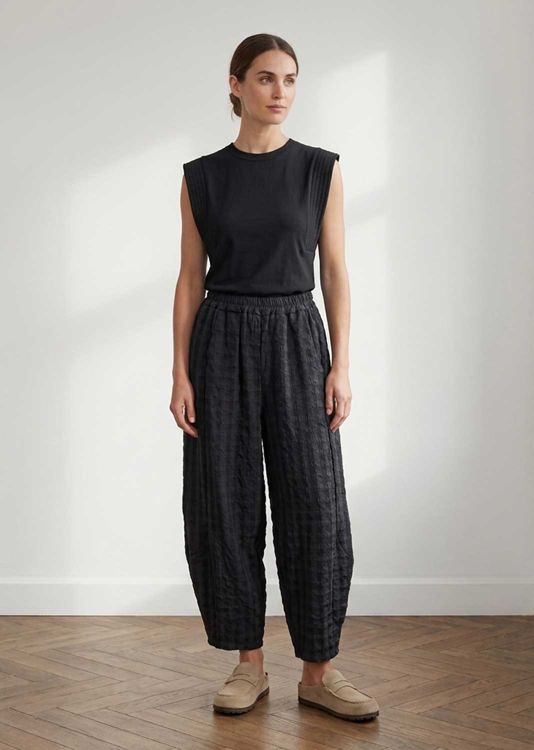 A woman stands in a minimalist room with a white wall and wood floor, wearing a sleeveless black top and dark charcoal patterned wide-leg pants with beige loafers.