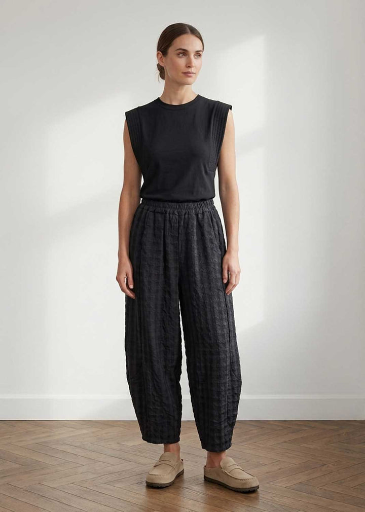A woman stands in a minimalist room with a white wall and wood floor, wearing a sleeveless black top and dark charcoal patterned wide-leg pants with beige loafers.
