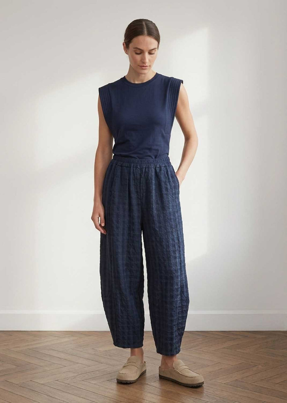 A woman stands in a minimalist room with a white wall and wood floor, wearing a sleeveless navy top and matching navy textured wide-leg pants with beige loafers.