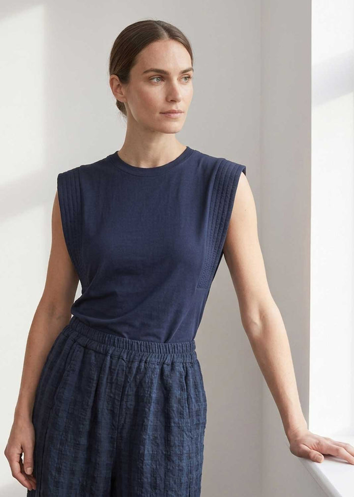 A woman wearing a sleeveless navy blue top and matching textured navy pants, standing in a brightly lit indoor setting.