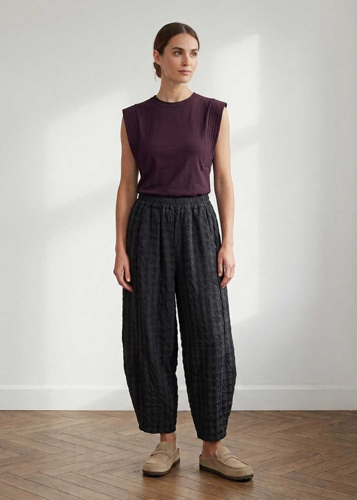 A woman stands in a minimalist room with a white wall and wood floor, wearing a sleeveless dark plum top and dark gray patterned wide-leg pants with beige loafers.
