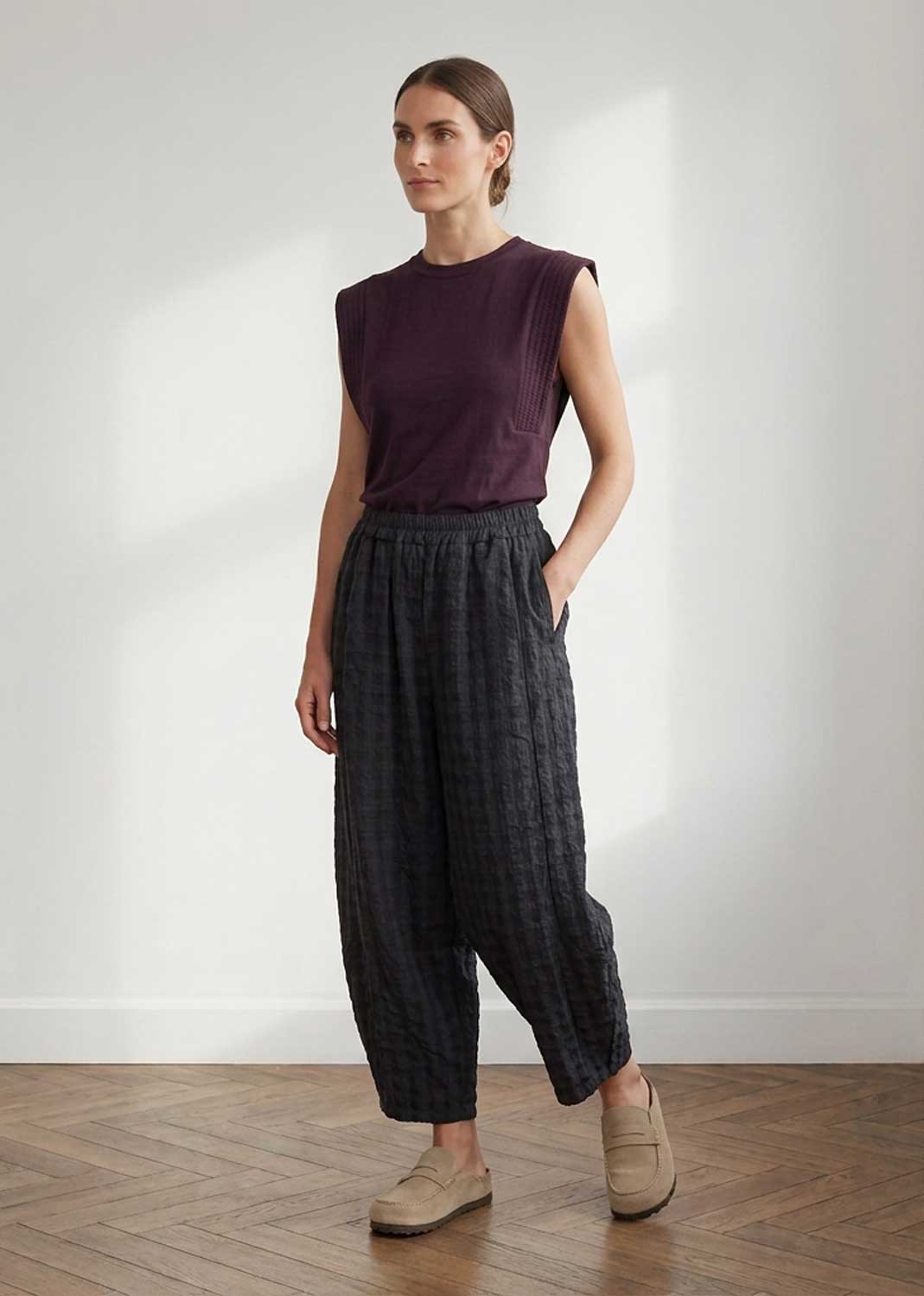 A woman modeling a sleeveless dark plum top and dark charcoal plaid wide-leg pants with beige loafers, standing in a minimalist room with a white wall and wooden floor.