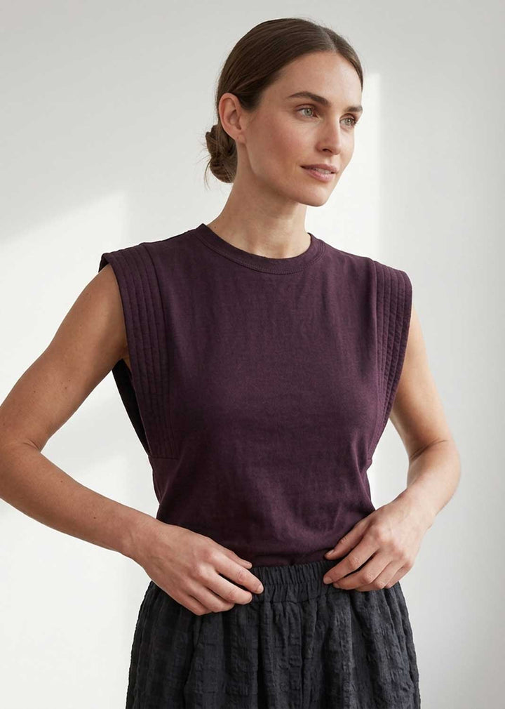 A woman wearing a sleeveless crew-neck knit top in deep plum purple, paired with a dark textured skirt, standing against a light gray background.