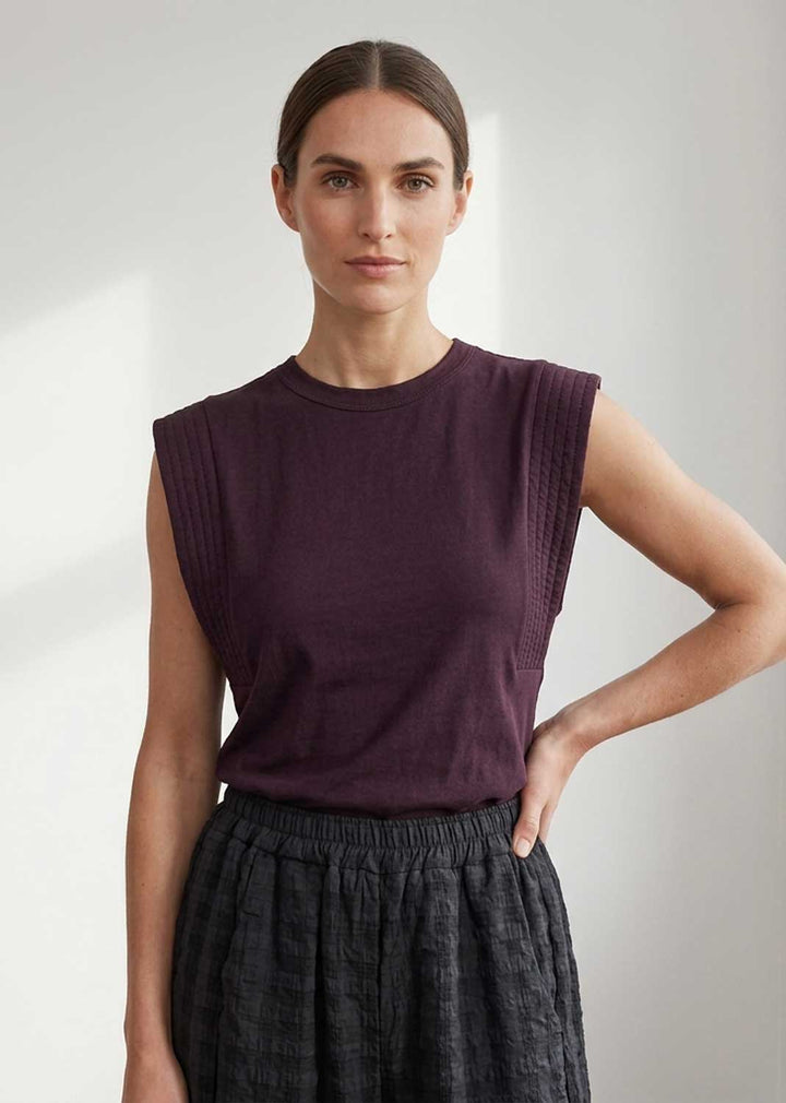 A woman modeling a sleeveless crewneck top in deep plum purple, paired with a dark textured skirt, against a neutral light gray background.