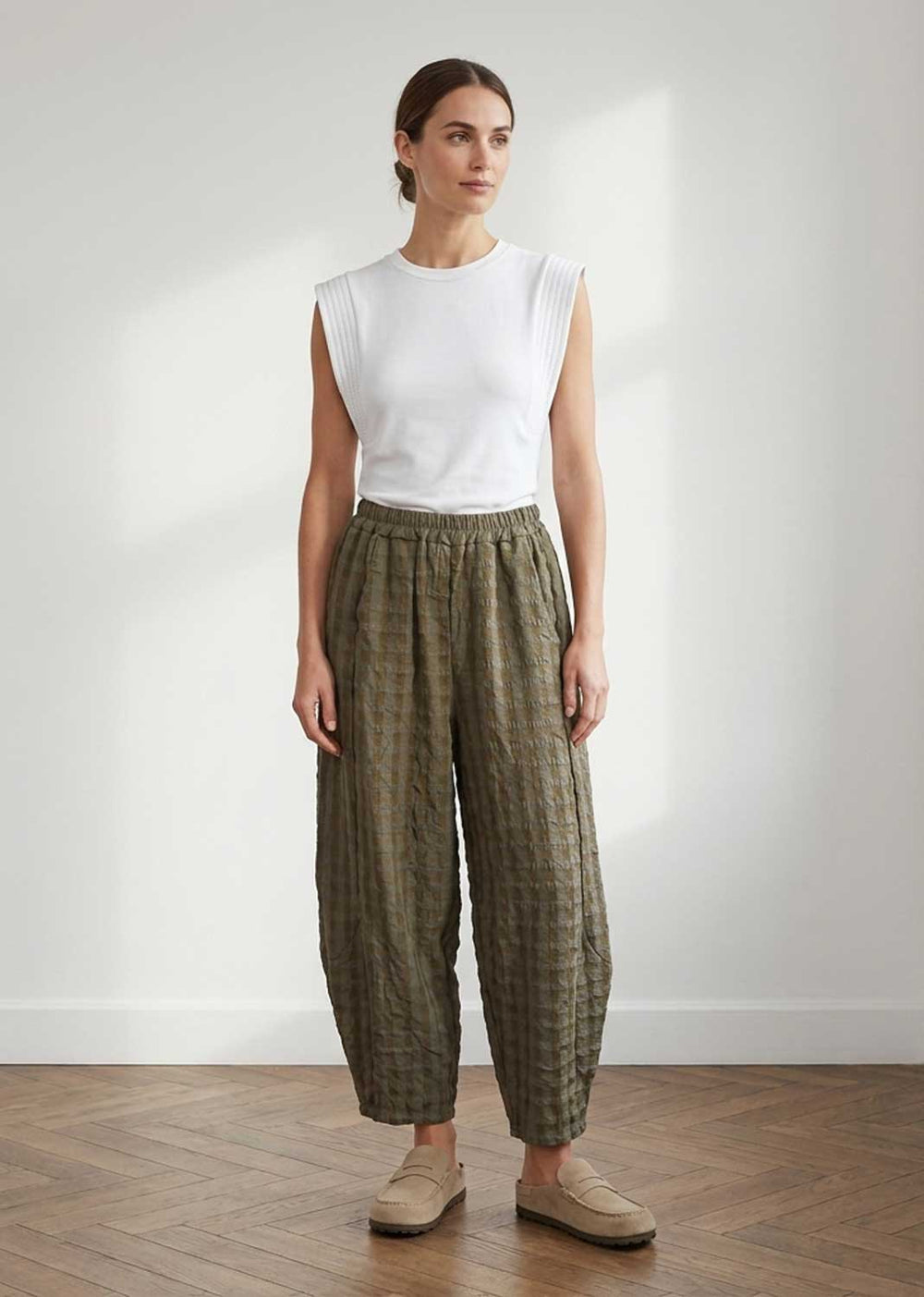 A woman stands in a bright room wearing a white sleeveless top and loose olive green patterned trousers with beige loafers.