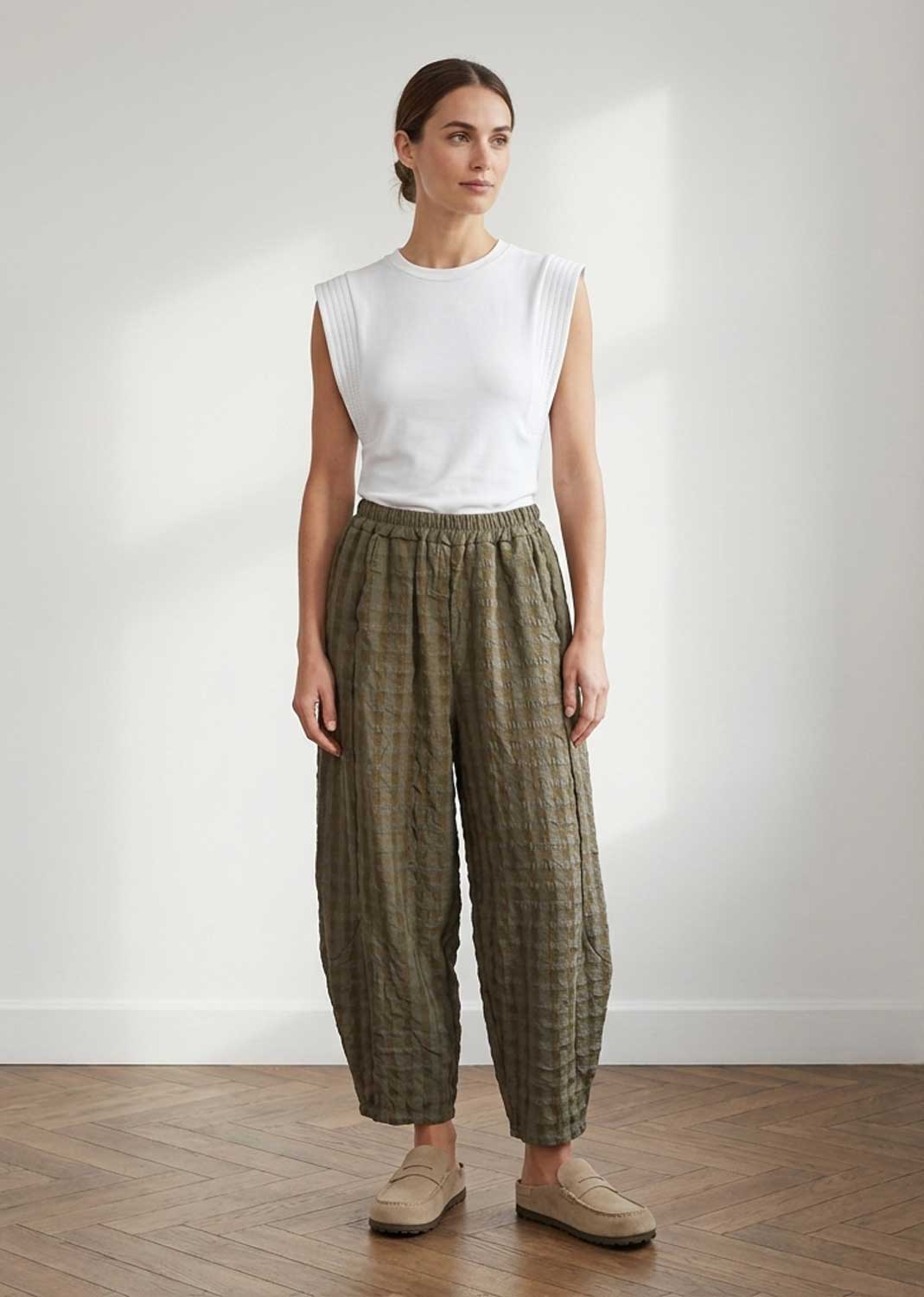 A woman stands in a bright room wearing a white sleeveless top and loose olive green patterned trousers with beige loafers.
