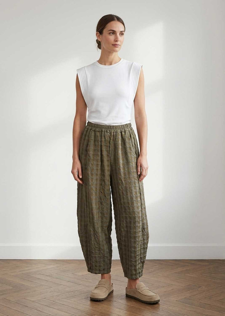 A woman stands in a bright room wearing a white sleeveless top and loose olive green patterned trousers with beige loafers.