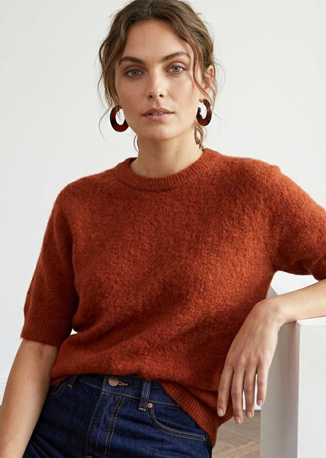 A woman wearing a short-sleeve crewneck knit sweater in a warm rust orange color, paired with dark blue jeans and large dark red hoop earrings, posed against a clean white background.