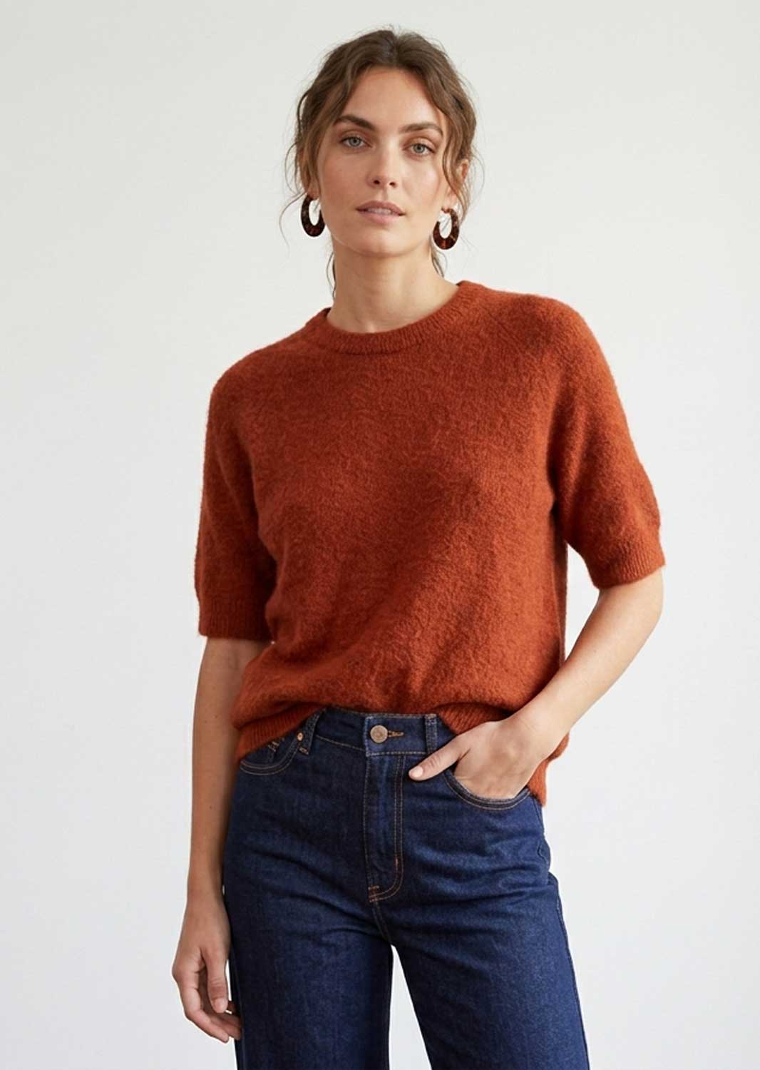 A woman wearing a short-sleeve, crew-neck knit sweater in a warm rust orange color, paired with dark blue jeans, against a plain light gray background.
