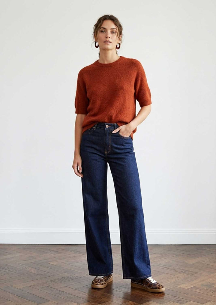 A woman stands in a studio wearing a rust-orange short-sleeve knit top, dark blue wide-leg jeans, and brown leather shoes against a white wall and wood floor.