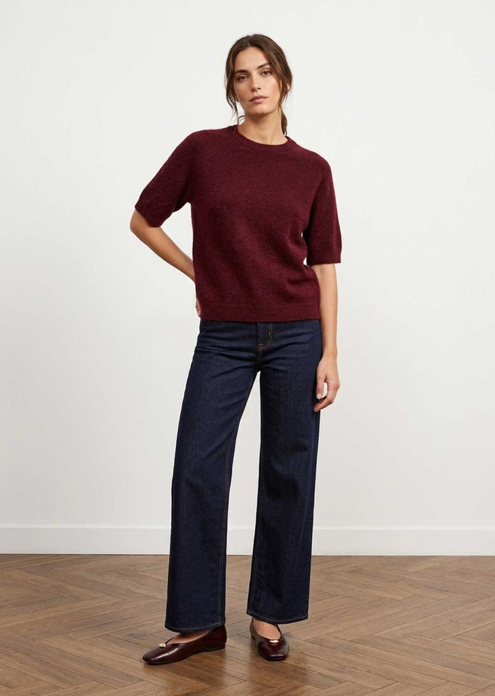 A woman modeling a short-sleeve crewneck knit sweater in burgundy, paired with dark blue wide-leg jeans and dark brown leather loafers, standing against a white wall on a wood floor.
