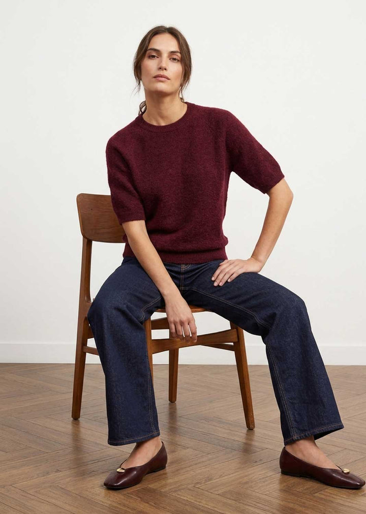 A woman sits on a wooden chair wearing a short-sleeve crewneck knit sweater in burgundy, dark wide-leg jeans, and dark brown leather loafers against a white wall and wood floor.