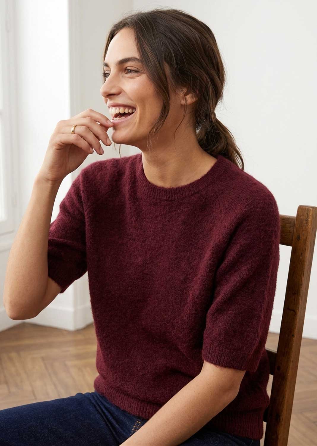 A woman laughing while wearing a short-sleeve crewneck knit sweater in burgundy, seated on a wooden chair in a bright indoor setting.